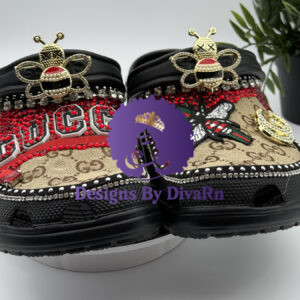 A pair of black Crocs heavily customized in a Gucci-inspired theme, featuring red rhinestones, logo-printed fabric, and large, decorative bee charms.