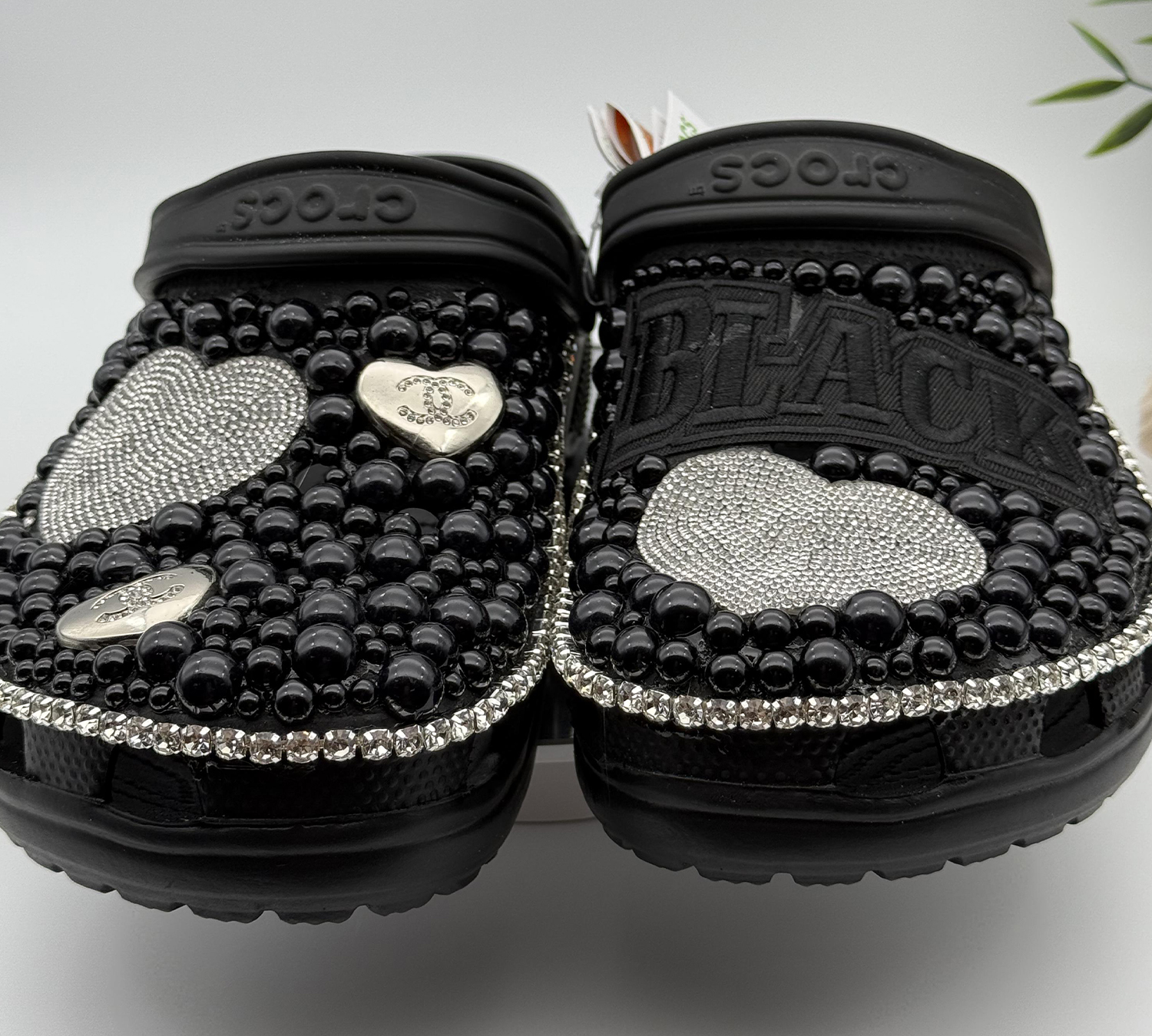 A pair of customized black Crocs, heavily embellished with black pearls, rhinestone trim, and silver rhinestone hearts.
