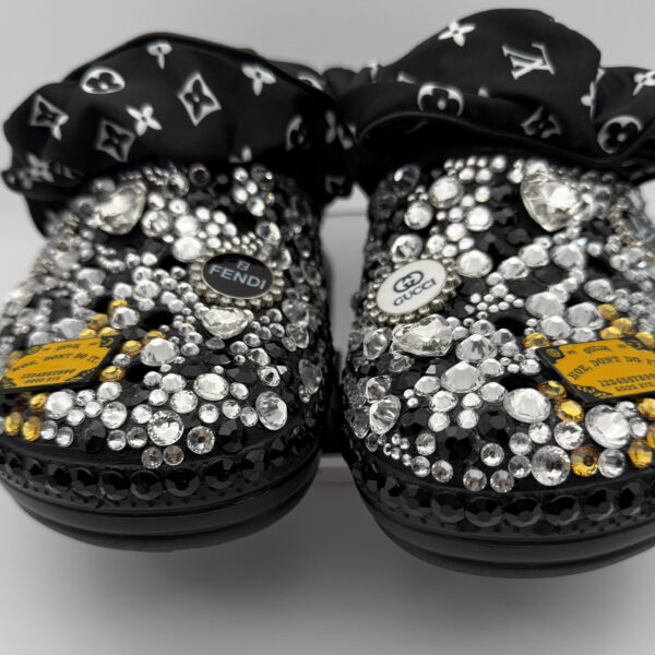 A pair of black clogs covered in clear, black, and iridescent rhinestones and featuring a Prada-inspired logo charm.