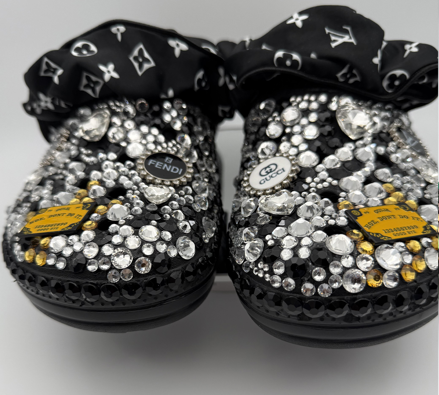 BLACRYCR-012 A pair of black clogs covered in clear, black, and iridescent rhinestones and featuring a Prada-inspired logo charm.