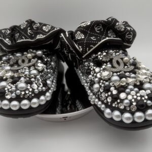 A pair of black custom clogs heavily embellished with black, silver, and grey pearls, clear rhinestones, and designer-inspired charms, finished with a black and white patterned fabric strap.