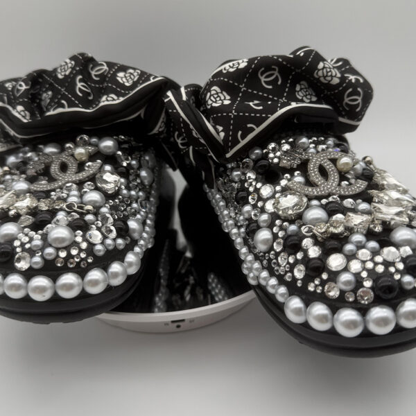 A pair of black custom clogs heavily embellished with black, silver, and grey pearls, clear rhinestones, and designer-inspired charms, finished with a black and white patterned fabric strap.