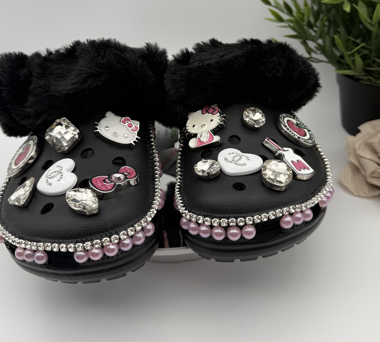 A pair of black, fur-lined clogs decorated with Hello Kitty charms, rhinestones, and pink pearls.