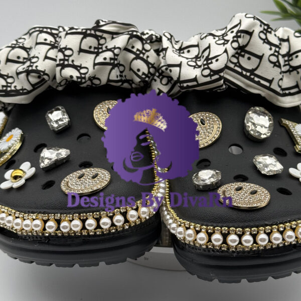 A pair of customized black Crocs featuring pearl and gold trim, designer-inspired scrunchie cuffs, and rhinestone smiley face and daisy charms.