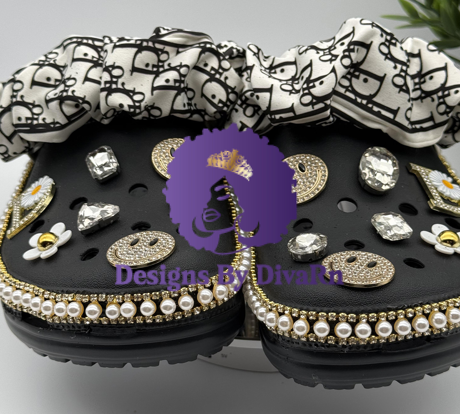 A pair of customized black Crocs featuring pearl and gold trim, designer-inspired scrunchie cuffs, and rhinestone smiley face and daisy charms.