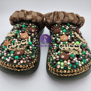 A pair of green custom clogs with designer-inspired teddy bear charms, a gold logo charm, green and gold rhinestones, and a monogrammed ruffled fabric strap.