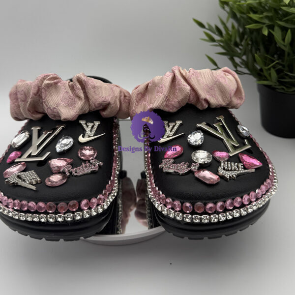A pair of black custom clogs with a pink ruffled fabric strap, decorated with silver designer-inspired charms, and pink and clear rhinestones.