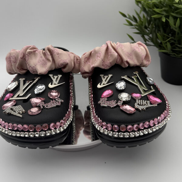 A pair of custom black clogs adorned with pink and clear rhinestones, designer-inspired charms, and a pink ruffled fabric strap.
