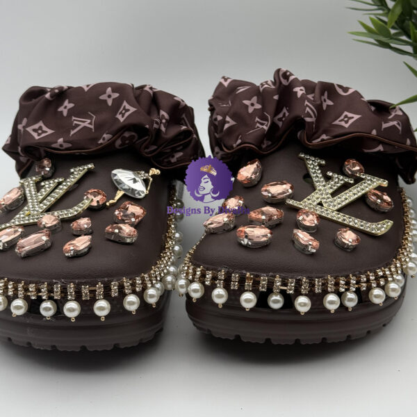 A pair of custom-decorated brown clogs by DesignsByDivaRn, displayed on a white surface with a small plant in the background. The clogs are embellished with blush-pink crystals, a dangling pearl and rhinestone trim, large sparkling initial charms, and a brown monogram-print scrunchie strap.