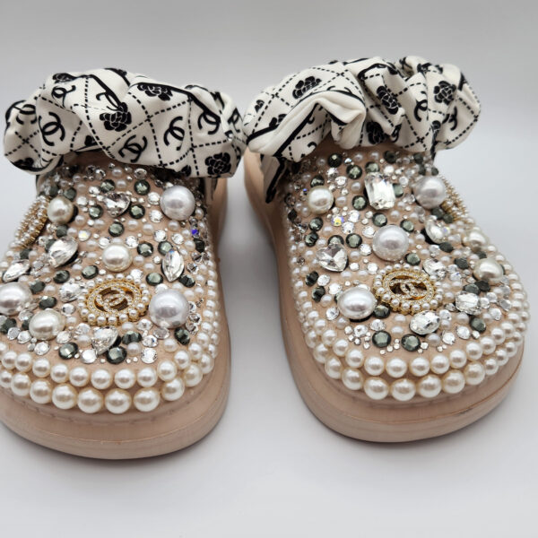 A pair of beige clogs beautifully customized by DesignsByDivaRn, covered in an elegant arrangement of pearls and clear crystals. The shoes feature gold-tone designer-inspired charms and are finished with a stylish black and white patterned scrunchie strap, shown on a white background.