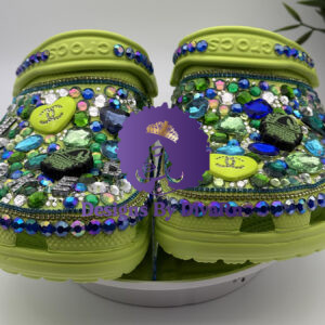 A front-facing shot of heavily customized lime green Crocs. The shoes are covered in a dense layer of green, blue, and iridescent rhinestones and adorned with charms, including a heart and a hand grenade. They are displayed on a white stand against a white background.