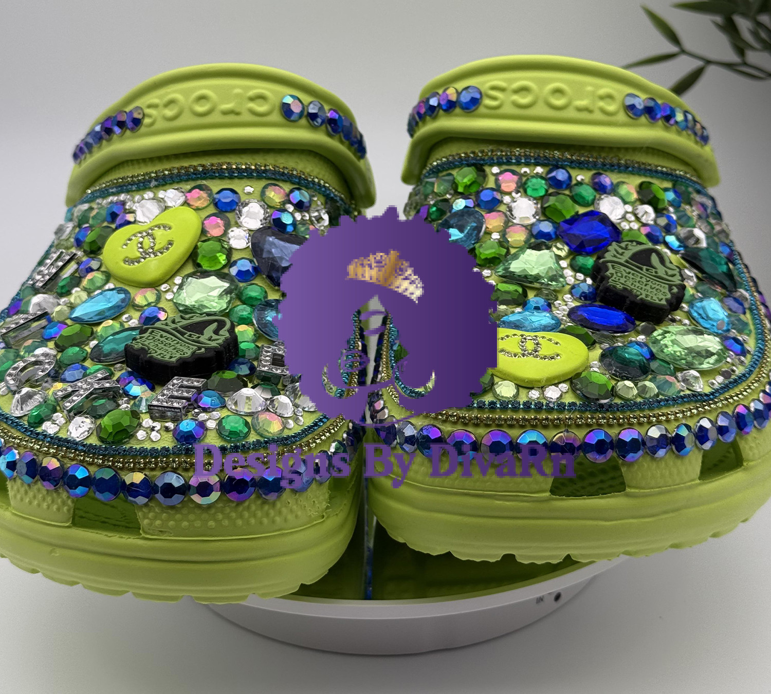 A front-facing shot of heavily customized lime green Crocs. The shoes are covered in a dense layer of green, blue, and iridescent rhinestones and adorned with charms, including a heart and a hand grenade. They are displayed on a white stand against a white background.