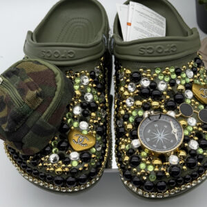 A pair of custom olive green clogs by DesignsByDivaRn, heavily decorated with black, gold, and green gems. One shoe features a miniature camouflage zippered pouch, while the other is adorned with a large compass charm.