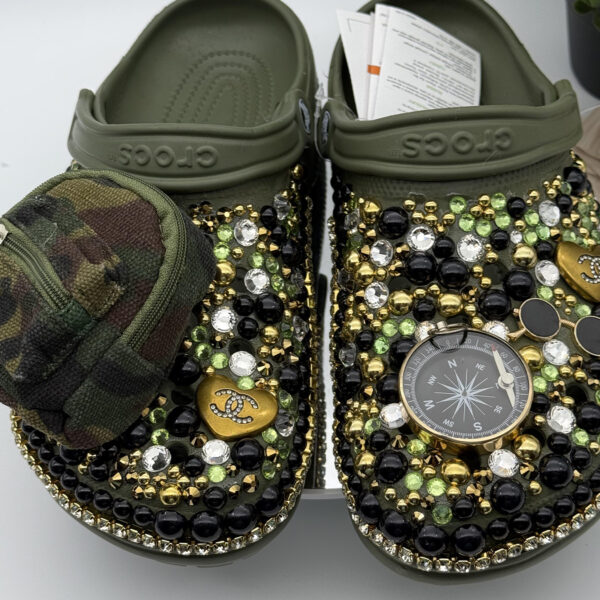 A pair of custom olive green clogs by DesignsByDivaRn, heavily decorated with black, gold, and green gems. One shoe features a miniature camouflage zippered pouch, while the other is adorned with a large compass charm.