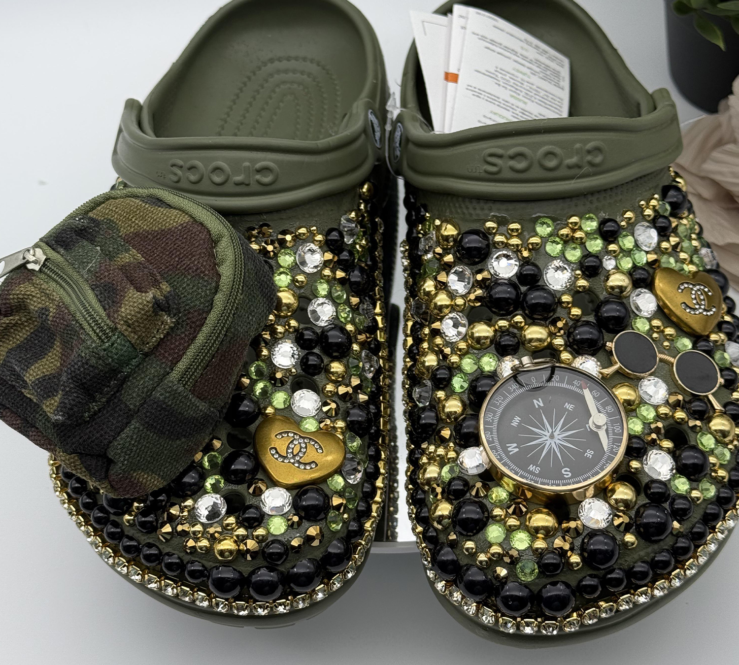 A pair of custom olive green clogs by DesignsByDivaRn, heavily decorated with black, gold, and green gems. One shoe features a miniature camouflage zippered pouch, while the other is adorned with a large compass charm.