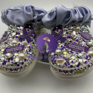 A pair of custom clogs covered in light pink faux fur with a rhinestone-trimmed toe. The clogs are decorated with various charms, including pearls, a round blue rhinestone charm, pink Chanel-inspired logos, and a prominent slogan charm that reads 