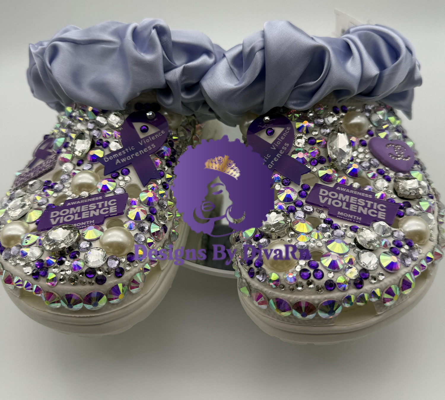 A pair of custom clogs covered in light pink faux fur with a rhinestone-trimmed toe. The clogs are decorated with various charms, including pearls, a round blue rhinestone charm, pink Chanel-inspired logos, and a prominent slogan charm that reads 