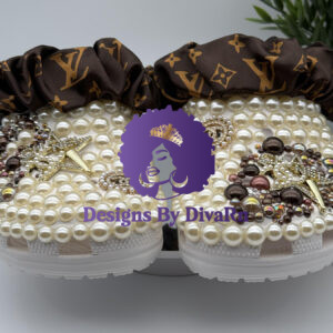 A pair of luxury custom white clogs by Designs by DivaRN, completely covered in cream pearls and adorned with gold pearl-encrusted star charms, rhinestone Chanel-inspired logos, and clusters of brown gems, finished with a brown Louis Vuitton-inspired monogram silk scrunchie on the strap.