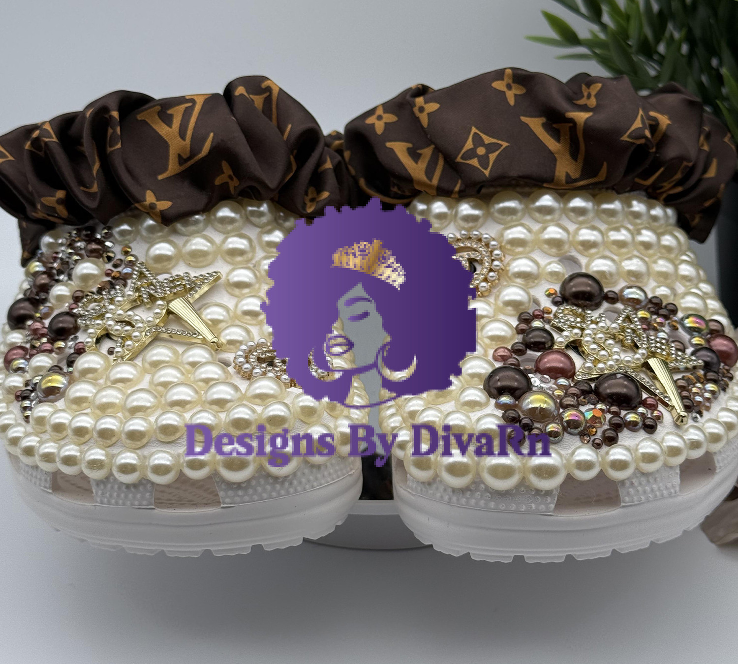 A pair of luxury custom white clogs by Designs by DivaRN, completely covered in cream pearls and adorned with gold pearl-encrusted star charms, rhinestone Chanel-inspired logos, and clusters of brown gems, finished with a brown Louis Vuitton-inspired monogram silk scrunchie on the strap.