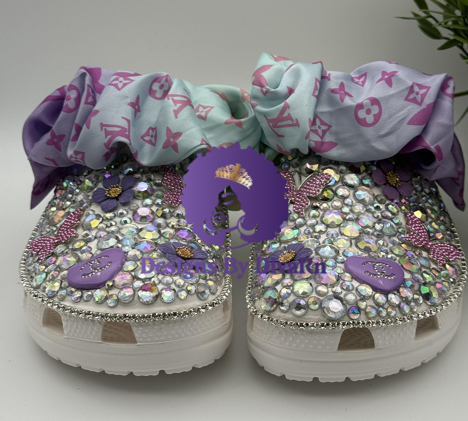 A pair of custom white clogs by Designs by DivaRN, lavishly covered in iridescent rhinestones, purple flowers, pink butterfly charms, and designer-inspired purple heart charms, with a pastel purple and mint monogrammed silk scarf tied around the strap.