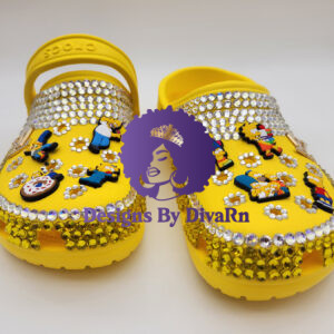 A pair of custom-designed yellow clogs by Designs By DivaRn, beautifully hand-bedazzled with clear and yellow rhinestones and decorated with colorful Jibbitz charms of The Simpsons characters, shown against a clean white background.