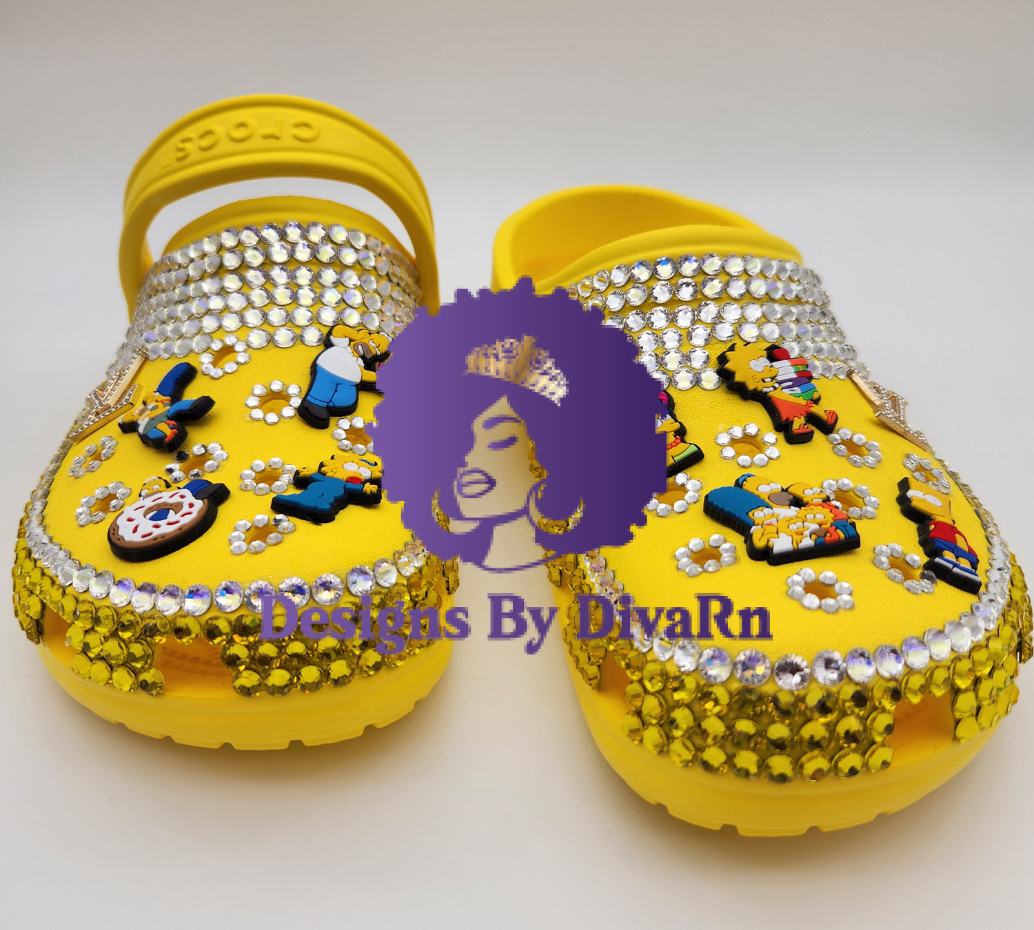 A pair of custom-designed yellow clogs by Designs By DivaRn, beautifully hand-bedazzled with clear and yellow rhinestones and decorated with colorful Jibbitz charms of The Simpsons characters, shown against a clean white background.