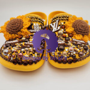A detailed front-view photograph of a pair of custom-designed yellow clogs by Designs By DivaRn. The shoes are lavishly embellished with hand-crocheted sunflowers, a dense mosaic of brown, amber, and clear rhinestones, iridescent pearls, and luxury-inspired charms including Givenchy, Louis Vuitton, and Chanel logos, all set against a clean white studio background.