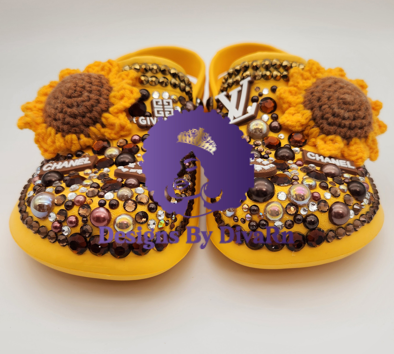 A detailed front-view photograph of a pair of custom-designed yellow clogs by Designs By DivaRn. The shoes are lavishly embellished with hand-crocheted sunflowers, a dense mosaic of brown, amber, and clear rhinestones, iridescent pearls, and luxury-inspired charms including Givenchy, Louis Vuitton, and Chanel logos, all set against a clean white studio background.