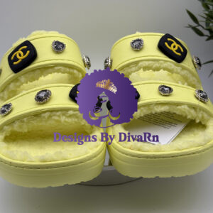 A pair of pastel yellow, dual-strap slides with a cozy shearling lining, hand-decorated by Designs By DivaRn with black and gold CC-style charms and heart-shaped gemstone jewels, shown on a white surface.