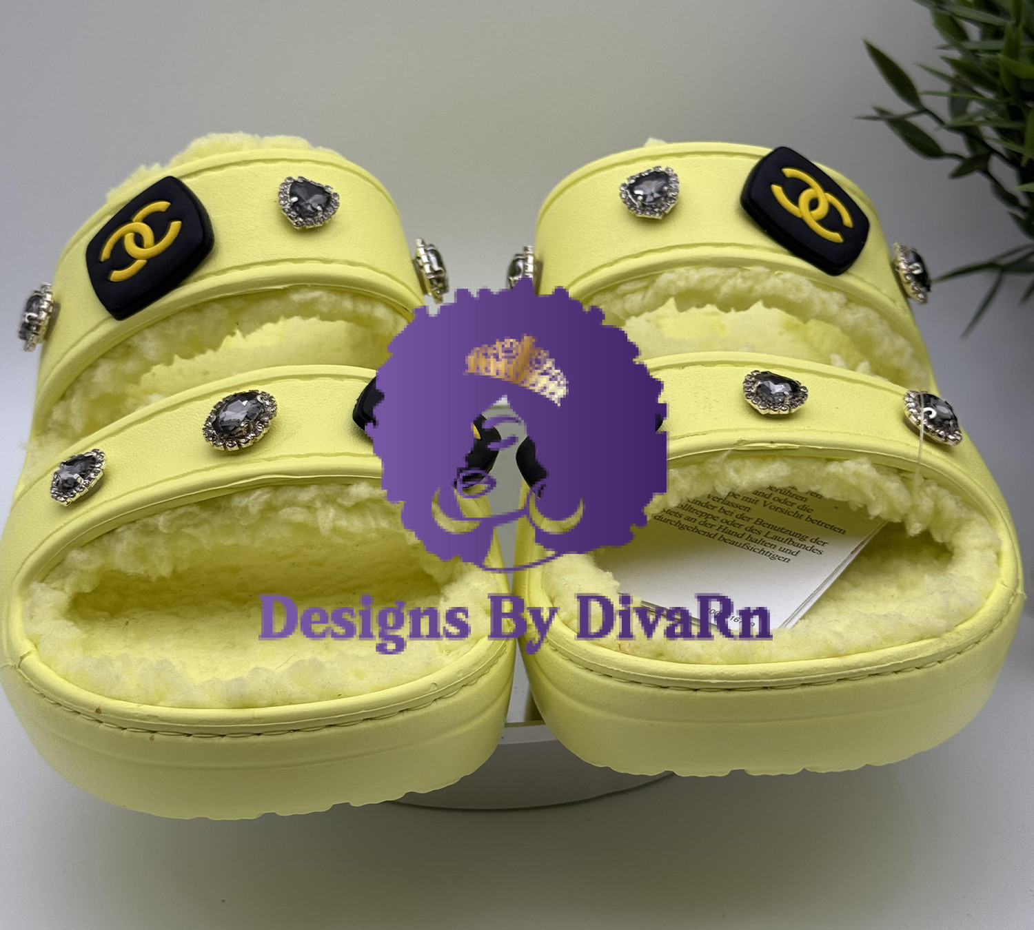 YELYELCR-077 A pair of pastel yellow, dual-strap slides with a cozy shearling lining, hand-decorated by Designs By DivaRn with black and gold CC-style charms and heart-shaped gemstone jewels, shown on a white surface.